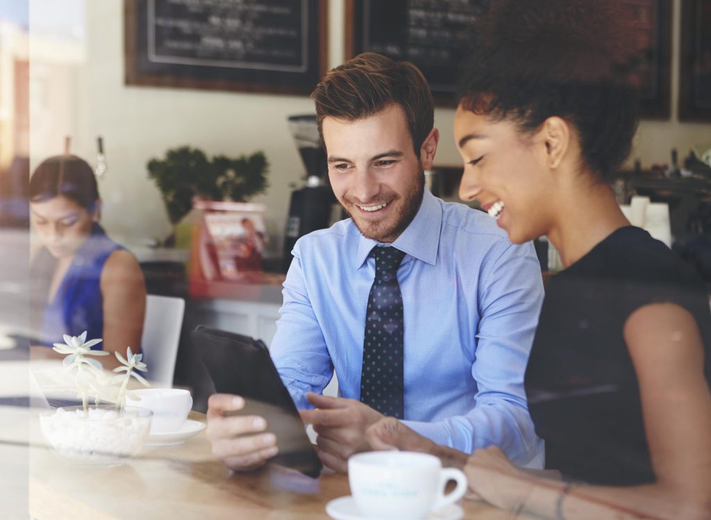 Coworkers share a laugh while looking at a tablet in a coffee shop.